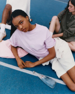 A young person with braided hair, dressed in a light purple shirt and white shorts, reclines on a blue surface. Another person sits nearby, legs partially visible. A glass bottle rests on the ground. The composition conveys a relaxed, casual atmosphere, with clean lines and a contemporary aesthetic, highlighting a sense of tranquility and modern style.
