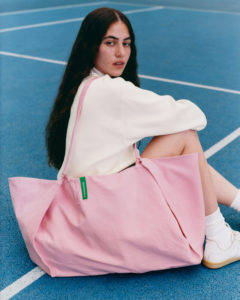 A person with long, dark hair sits on a blue court, wearing a white long-sleeve outfit and white sneakers. They carry a large, soft-looking pink bag over their shoulder, featuring a green tag. Their relaxed pose and neutral expression suggest a casual and stylish moment, blending fashion with a sporty, outdoor setting.