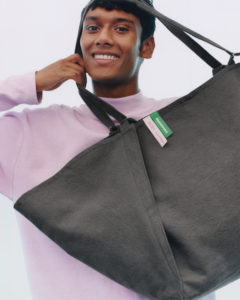 A smiling person is holding a large, dark-colored tote bag. The person is wearing a light purple sweater, and the background is plain, enhancing the casual and cheerful vibe. The tote bag features simple lines and a rectangular shape, highlighted by a small green tag. The overall scene conveys a sense of modern style and practicality.