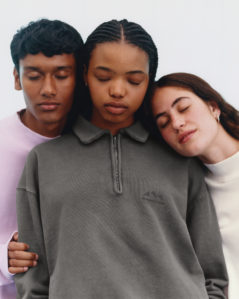Three young individuals stand closely with their eyes gently closed, creating a tranquil and harmonious scene. The person in the center wears a dark zip-up sweater, while the two on either side are in lighter tops. Their postures convey a sense of unity and calm, with one leaning their head on the other's shoulder, adding an intimate, peaceful touch.