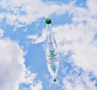 A clear bottle stands upright in the center, with the sky and clouds reflecting below. The water surface creates a rippled effect, enhancing the reflection's symmetry. The bottle has a green cap and is filled with clear liquid. Surrounding the reflection are soft white clouds, giving a tranquil and refreshing ambiance against a light blue background.