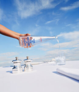 A hand pours clear liquid from a tall, transparent bottle into a glass. The backdrop features a bright blue sky with scattered clouds, creating a sense of openness. In the foreground, a set of silver salt and pepper shakers rest on a pristine white surface, alongside neatly arranged white napkins and cutlery.