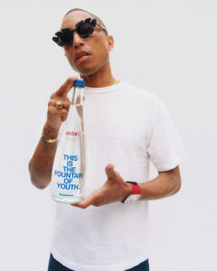 A stylish individual wearing unique sunglasses and a white shirt holds a clear water bottle. Their hands display several rings, a tattoo, and a red wristwatch. The person exudes confidence and modern flair against a simple, clean background, drawing attention to both their fashion choices and the prominently featured bottle in their hand.