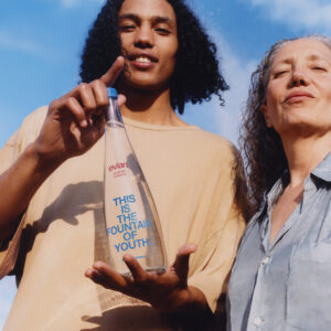 Two individuals stand closely under a clear blue sky. The younger person on the left holds a bottle, pointing at it with a smile. The older individual on the right looks ahead with a calm expression. Both have curly hair and are dressed casually. The scene conveys a sense of connection and vitality through their expressions and postures.
