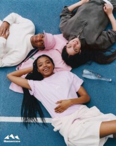 Three people are joyfully lying on a blue surface, forming a connected trio. One wears a pink cap and white sweatshirt, another is in a gray hoodie, and the third sports a light purple shirt with braided hair. They appear relaxed and content, forming a harmonious scene with casual, colorful attire. A clear bottle lies nearby, adding to the relaxed atmosphere.