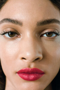 A close-up of a person's face showcasing a smooth complexion and perfectly groomed eyebrows. The focus is on striking, vivid red lips contrasted against natural skin tones. Eyes are accentuated with long lashes, giving a captivating look. Subtle shadows highlight facial features, creating a balanced and harmonious appearance that draws attention to the details.
