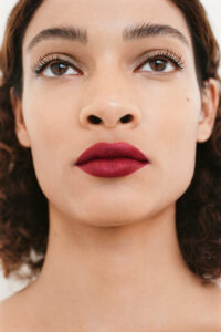 A person with curly brown hair is depicted with a focus on their face. Their expression is neutral, and they wear bold, matte red lipstick. The skin appears smooth and even-toned. The eyebrows are well-groomed, and the eyes are accentuated with long, dark eyelashes. Soft lighting enhances their features against a neutral background.
