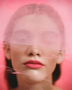 A profile showcases a woman's face partially obscured by a misty or frosted effect. Her lips are bright red and prominent against her fair complexion. The overall tone is pink, giving a dreamy, ethereal quality to the scene. The soft blending and partial concealment evoke a sense of mystery and artistic abstraction.