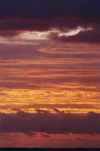 A serene view showcases a vibrant sky filled with varying hues of orange, red, and yellow as the sun sets. Streaks of clouds add dramatic contrast against the brightly illuminated horizon. The lower clouds appear darker, suggesting the closing of the day, creating a tranquil and picturesque scene that evokes a sense of peace and wonder.