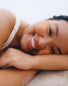 A person lies on their side, smiling softly. They have short, dark hair and wear a white shirt. Their hand rests near their face on a sandy surface, creating a relaxed and serene atmosphere. The natural lighting highlights their content expression, capturing a moment of tranquility and ease. The background is simple and unobtrusive.