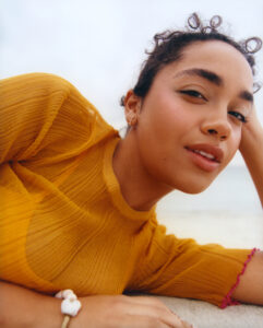 A person with short, curly hair is reclining with a relaxed expression. They are wearing a yellow ribbed top and a bracelet with a white detail on their wrist. The background is softly blurred, suggesting an outdoor setting with a light, natural ambiance, possibly by the sea. The overall mood is calm and serene.