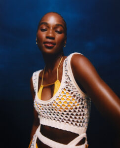 A confident individual poses against a dark, moody background. They are wearing a white, crochet-style top over a bright yellow garment, adding vibrant contrast. The lighting softly highlights their skin, accentuating their serene expression and poised demeanor. Subtle accessories adorn their ears, complementing the overall look and adding an elegant touch to the fashionable ensemble.
