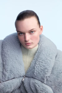 A person with light skin and short, slicked-back hair gazes directly forward. They wear a thick, textured, light gray coat with a high collar, partially zipped, over a beige turtleneck. The background is a soft, pale blue, giving a clean and minimalist appearance. The overall mood suggests modernity and minimalism, accentuating the person's calm and composed demeanor.