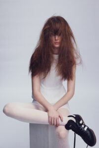 A person with voluminous, long brown hair partially covering their face sits with legs crossed. They wear a sleek white clothing ensemble, including tights. A sturdy, block-like structure supports them. Their feet are adorned with stylish black sneakers featuring white stripes. The backdrop is a smooth, neutral tone, emphasizing the subject's striking presence and unique style.
