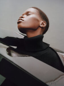 A person faces upward with eyes closed, exuding calm and poise. They wear a black turtleneck sweater, complementing their short hair. The lighting highlights their facial features, casting soft shadows. Reflections surround them, adding layers to the composition, while a neutral background contrasts with the subject’s dark clothing, emphasizing their serene demeanor and artistic pose.