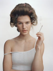 A woman with an elegant updo, adorned with curlers, holds a cigarette. Her white, delicate top features subtle patterns. Visible smoke adds an ethereal touch, enhancing the serene and introspective mood. Her expression is calm and confident, conveying a sense of quiet contemplation amid the softly lit, minimalist backdrop. The overall atmosphere is classic and poised.