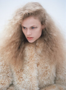 A person with striking, voluminous blond hair gazes intensely to the side. Their complexion is fair, with bold, artistic eyeliner accentuating their eyes. They are wearing a textured, fluffy coat that matches their hair color, enhancing the overall visual impact. The background is plain and muted, directing focus entirely on the individual's expressive features and dramatic appearance.