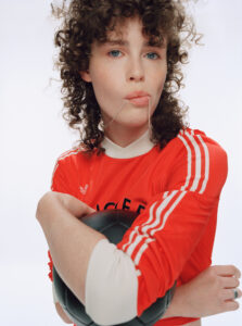 A person with curly hair gazes forward, holding a black ball close to their body. They wear a bright red, long-sleeved athletic top with white stripes on the sleeves. A silver necklace is held in their mouth, adding a playful, contemplative element. The background is a soft, neutral tone, highlighting the person’s features and clothing.