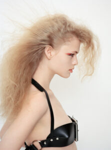 A person with voluminous light hair styled in a wild, teased manner gazes intently to the side. The striking makeup features bold red liner accentuating the eyes. The individual wears a unique black leather outfit with straps and metal detailing, creating a modern and edgy fashion statement. The background is plain and soft, highlighting the subject.