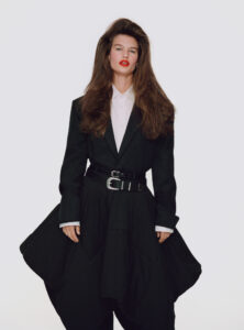 A person stands confidently, dressed in a stylish black ensemble with an oversized coat and voluminous trousers. The outfit features a sleek white shirt underneath and is cinched at the waist with a wide belt. The individual's long, voluminous hair complements their striking red lipstick, blending elegance and modern fashion in a minimalistic setting.