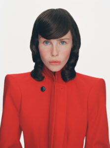 A person with striking blue eyes gazes directly ahead, wearing a bold red coat with a high collar and a single black button. The hairstyle is smooth with soft curls framing the face. The expression is serious, set against a plain white background, highlighting the vivid contrast between the attire and the backdrop.