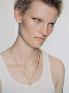 An individual with short, light hair and blue eyes gazes off to the side with a serious expression. Their skin appears fair with freckles. They wear bold eyeliner, emphasizing the eyes, and a white tank top. A delicate gold necklace with a pendant rests around their neck, standing out against the simple background.
