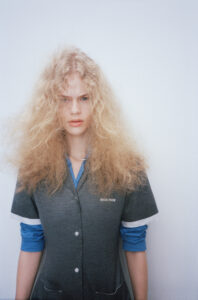 A person with voluminous, curly blonde hair stands against a plain background. They wear a layered outfit with a grey button-down cardigan and a blue top underneath, the cuffs rolled up. The expression suggests a serious or contemplative mood, and the absence of visible surroundings creates a stark, minimalist atmosphere, emphasizing their distinctive hairstyle and clothing.