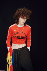 A person with curly hair is wearing a red cropped long-sleeve shirt paired with black pants. A simple chain necklace adorns the neck, and there’s a visible tattoo on the lower abdomen. A colorful garment is draped over the forearm. The overall look is casual yet edgy, set against a plain, dark background.