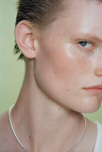 A close-up view of a person with fair skin and small freckles, showcasing the side of the face with slicked-back hair. Only one blue eye is visible, capturing a focused expression. The background is a muted light green, and the person is wearing a thin silver necklace and a white sleeveless shirt. The skin appears smooth and slightly shiny.