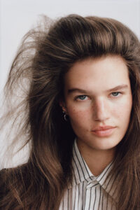 A woman with striking features gazes directly ahead. She has voluminous, flowing brown hair framed around her face, and her complexion is highlighted with freckles. Her expression is calm yet intense, and she wears small hoop earrings. A collared shirt with vertical stripes adds a touch of sophistication to her overall appearance. The background is neutral.