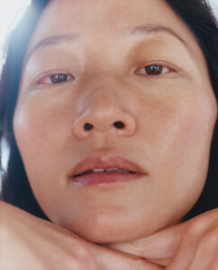 A close-up depicts a serene expression with relaxed features. The light highlights smooth skin with subtle natural tones. Eyes appear calm and contemplative, while the soft focus adds a gentle feel. Dark hair frames the face, and hands are elegantly positioned under the chin, offering a sense of tranquility and introspection.