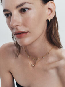 A person gazes thoughtfully to the side. They have subtle makeup, emphasizing natural features. Gold hoop earrings adorn their ears, while layered necklaces of fine gold chains with small, sparkling pendants grace their neck. The background is softly blurred, focusing attention on the serene expression and elegant jewelry. Light illuminates the skin, creating a gentle, radiant effect.