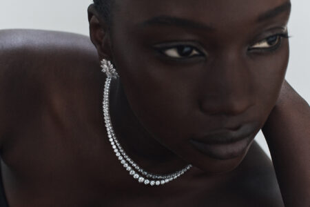 A person adorned in elegant jewelry poses gracefully, displaying a sparkling necklace and an intricate earring. The lighting enhances the gleaming jewels and the soft texture of the skin, creating an atmosphere of luxury and sophistication. The focus captures the poised expression, emphasizing the delicate design and craftsmanship of the accessories, evoking a sense of refined elegance.