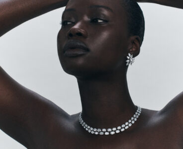 A person with a serene expression gazes upwards, arms gently raised over their head. They wear a stunning diamond necklace with an elegant double strand design. Complementing this, their earrings feature intricate floral-shaped clusters, adding a touch of sophistication. The minimalist, neutral background enhances the elegance and focus on the jewelry and the subject's graceful pose.