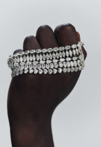 A hand with dark skin elegantly grasps a luxurious arrangement of sparkling diamond bracelets. Each bracelet features rows of clustered diamonds in various cuts, creating a dazzling and opulent effect. The contrast between the rich skin tone and the shimmering jewels highlights the exquisite craftsmanship and glamour of the jewelry, set against a minimalistic light background.