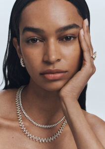 A woman with smooth skin and long, dark hair gazes forward with a calm expression, resting her cheek on her hand. She wears elegant jewelry: a pair of earrings, a ring with intricate details, and two layered necklaces, each adorned with sparkling diamonds, adding a touch of sophistication. Her nails are neatly manicured, complementing her overall polished appearance.