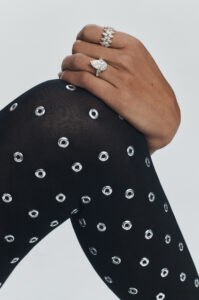 A hand rests elegantly on a black, patterned fabric adorned with silver grommets. The hand is adorned with two striking rings: one features a large pear-shaped stone, while the other is a layered band with multiple marquise-shaped stones. The contrasting textures and elements create a luxurious and sophisticated aesthetic, accentuating both jewelry and fabric.