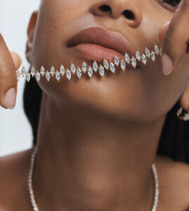 A close-up view features a person holding a delicate, sparkling diamond bracelet near their lips. The bracelet consists of marquise-cut diamonds, elegantly linked in a continuous line. Their lips are a natural shade, and they wear a necklace that complements the bracelet. The background is softly lit, creating a sense of elegance and sophistication.