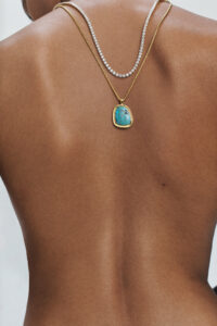 A bare back elegantly adorned with two necklaces: one featuring a row of small, sparkling stones, and the other showcasing a striking turquoise pendant set in gold. The necklaces rest gracefully, striking a balance between simplicity and luxury. The background is subtly blurred, drawing focus to the textures and colors of the skin and jewelry.