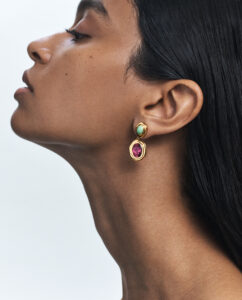 A woman is shown in profile, emphasizing her long neck and smooth skin. Her dark hair is sleek and tucked behind her ear, adorned with an elegant earring. The earring features a golden setting with a bright turquoise stone above a larger, vivid pink gem. The background is an understated, soft, pale color.