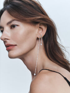 A woman gazes thoughtfully, highlighting her elegant profile. Her long brown hair cascades down, neatly tucked behind her ear. She wears striking, elongated drop earrings adorned with sparkling gems that dangle gracefully, catching the light. Her makeup is subtle, with a focus on natural tones, while her attire features a delicate, thin-strapped black top.