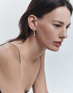 A woman with fair skin and dark hair gazes downward in a contemplative pose. She is wearing elegant, long, dangling earrings with multiple gemstones. Her attire is simple, featuring thin straps, which contrasts with the soft, light background that surrounds her. The subtle makeup enhances her natural features, adding to the poised and sophisticated essence of the scene.