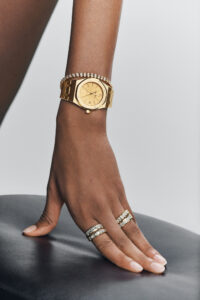 A hand with manicured nails is elegantly posed on a dark leather surface. On the wrist, there's a luxurious gold watch with a bejeweled bezel. The fingers are adorned with two stacked rings, each featuring sparkling stones. The overall tone is sophisticated and stylish, highlighting the shimmering jewelry against a clean, minimalist background.