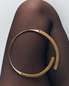Against a backdrop of dark, sheer fabric, a curved, gold-toned accessory glistens, partially studded with sparkling gems. The piece forms an incomplete circle, its ends elegantly tapered. Light reflects off its polished surface, enhancing the contrast between the smooth metallic texture and the soft, textured background, creating a sophisticated and luxurious visual composition.