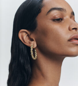 A person gazes serenely to the side, showcasing their elegant profile. Their sleek black hair enhances the clean, minimalistic aesthetic. A gold hoop earring, adorned with sparkling gemstones, complements the smooth skin tone. The overall composition exudes sophistication and timeless beauty, capturing a moment of understated glamour against a soft, neutral background.