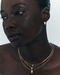 A person with an elegant and serene expression is shown wearing minimalistic jewelry. Three gold necklaces of varying lengths each hold a teardrop-shaped pendant, harmoniously complementing a single, delicate earring. The lighting softly highlights the contours of the face and accessories, creating a graceful and sophisticated ambiance. The color palette is warm and understated, enhancing the overall elegance.