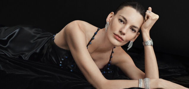 A woman reclines gracefully, wearing a sleek black satin dress adorned with small beads. Her elegant jewelry includes sparkling earrings, a watch, and a ring. Her pose is relaxed yet poised, with her head resting on one hand. Her expression is serene, set against a dark background that emphasizes her sophisticated appearance and timeless style.