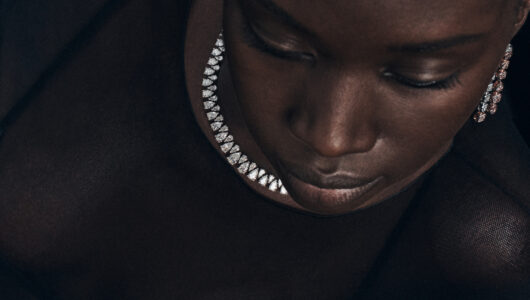 A close-up perspective highlights a woman's serene expression, emphasizing her closed eyes and slightly parted lips. She wears a sheer, dark garment adorned with a luminous, studded necklace. Her features are gentle and calm, enhancing the elegance of the sparkling jewelry around her neck and ears. The overall mood is one of quiet sophistication and introspection.