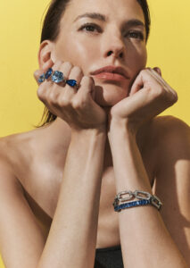 A woman with a contemplative expression rests her chin on her hands against a vibrant yellow background. She wears multiple rings adorned with large blue gemstones and a matching bracelet with silver links and blue accents. Her bare shoulders and natural makeup enhance the focus on the jewelry and the bright contrasting backdrop.