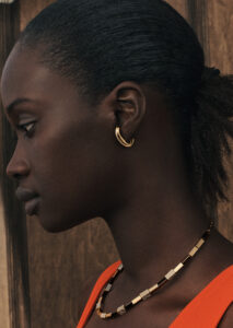 A woman is shown in profile, sporting a sleek hairstyle that pulls her hair back. She wears a golden hoop earring and a stylish, geometric necklace crafted with metallic and possibly encrusted segments. Her clothing is a bright, warm shade, contrasting subtly with the wooden background, which creates a natural and earthy aesthetic.